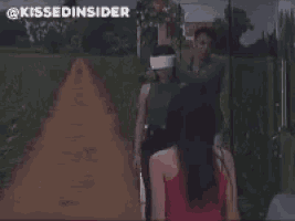 a group of people are standing on a dirt road with a man holding a blindfolded woman 's head .