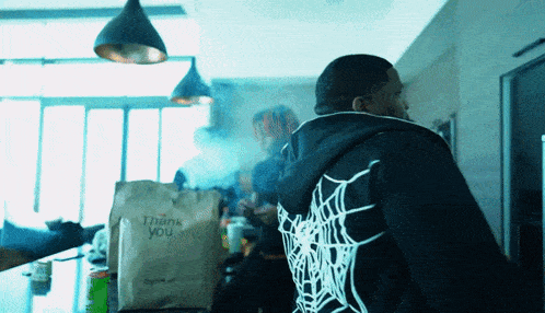 a man wearing a black hoodie with a spider web on the back is standing in front of a bag that says thank you