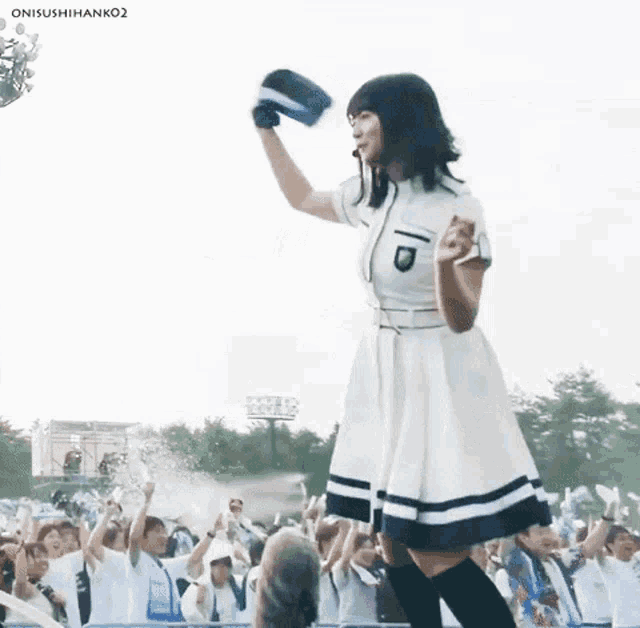 a woman in a white dress is standing in front of a crowd holding a baseball cap in her hand .