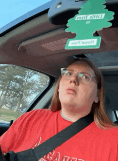 a woman wearing glasses and a red shirt is driving a car with a little trees air freshener on the windshield