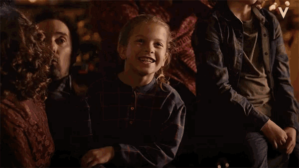 a little girl in a plaid shirt smiles while sitting in a group of people with the letter v in the corner