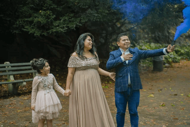 a man in a suit holds a blue smoke bomb in front of a pregnant woman and a little girl