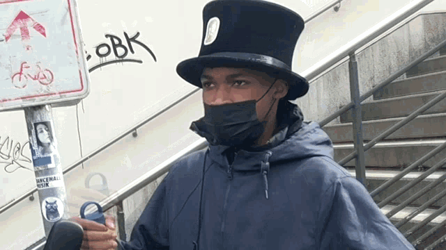 a man wearing a mask and a top hat with obk written on the wall behind him
