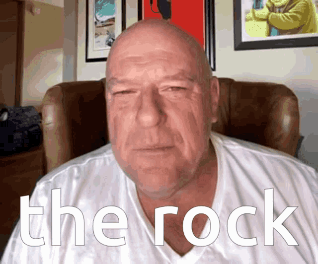 a bald man in a white shirt is sitting in a chair with the word the rock written on his shirt