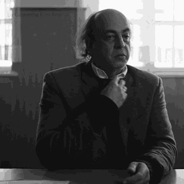 a man in a suit sits at a table in front of a window with la guardia film festival written on the bottom left