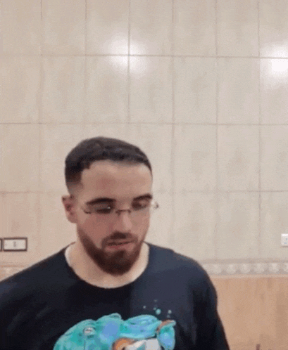 a man with a beard and glasses is standing in front of a tiled wall and looking at the camera .