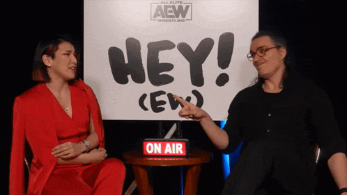 a man and a woman sit in front of a sign that says hey