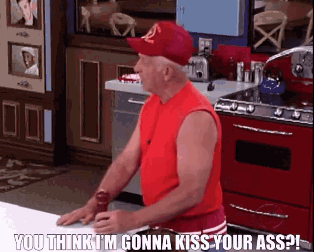 a man in a red shirt is sitting in a kitchen with the words " you think i 'm gonna kiss your ass " below him