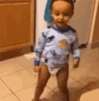 a little boy in a diaper is standing on a tiled floor in a kitchen .