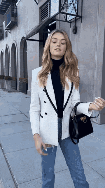 a woman wearing a black turtleneck and a white blazer is standing on a sidewalk
