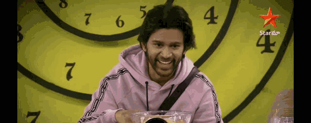 a man in a pink hoodie is smiling in front of a clock that says star