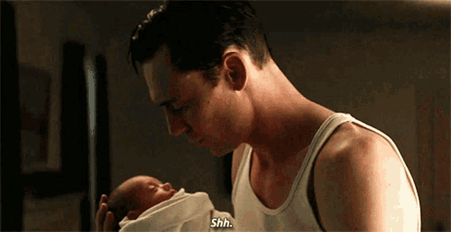 a man in a white tank top is holding a baby and says shh