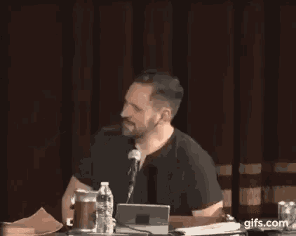 a man with a beard is sitting at a table with a laptop and a microphone while giving a speech .