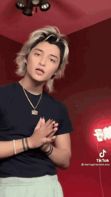 a man with blonde hair is wearing a black shirt and a necklace while standing in front of a red wall .
