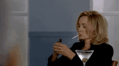 a woman is smoking a cigarette while sitting at a table with a martini .
