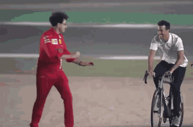 a man in a red suit is standing next to a man in a white shirt riding a bike on a race track .