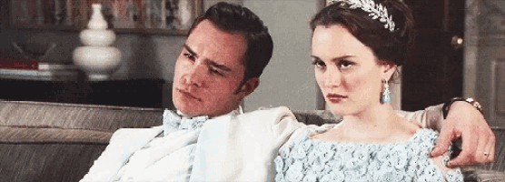 a man and a woman are sitting on a couch . the woman is wearing a blue dress and a headband .
