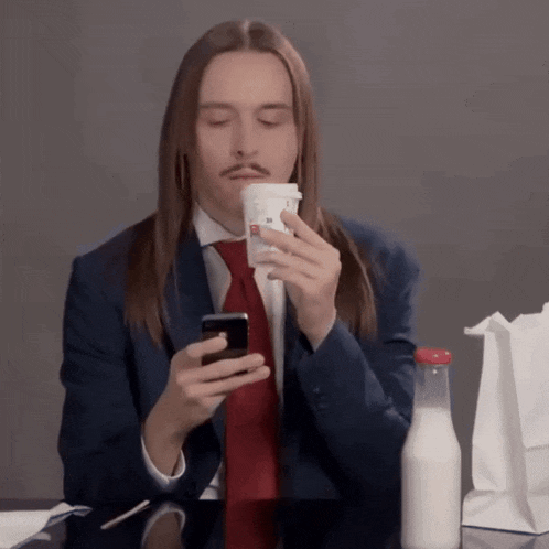a man with long hair is holding a cup of coffee and looking at his cell phone