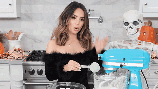 a woman standing next to a blue kitchenaid mixer with a skeleton in the background