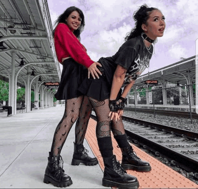 two women are posing for a picture at a train station . one of the women is wearing a red sweater .