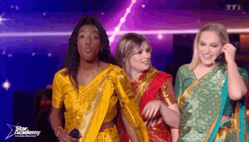 three women wearing saris are standing on a stage and one is holding a microphone .
