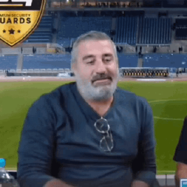 a man with a beard and glasses is sitting in front of a stadium with a premium security guards logo