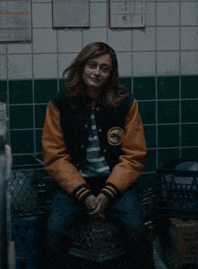 a woman in a varsity jacket sits on a trash can
