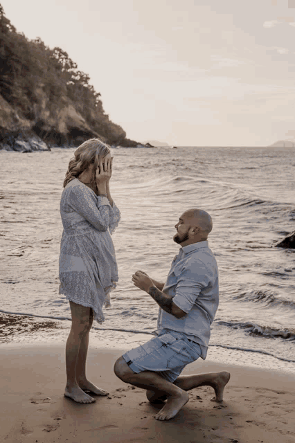a man kneels down to propose to a woman on a beach
