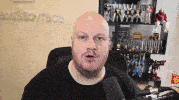 a bald man with a beard is talking into a microphone in front of a shelf full of toys .