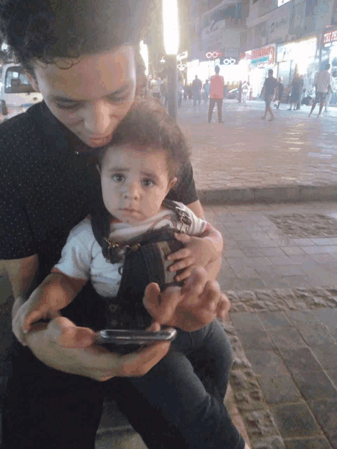 a man is holding a baby and looking at a cell phone in front of a store that says ' optical '