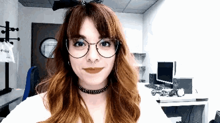 a woman wearing glasses and a choker looks at the camera in an office