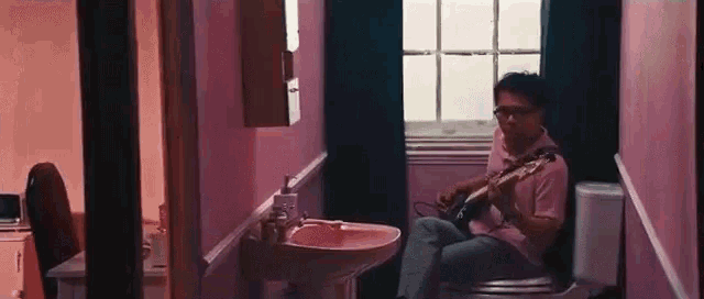 a man is playing a guitar while sitting on a toilet in a bathroom