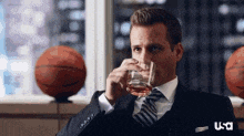 a man in a suit and tie is drinking a glass of whiskey while sitting in front of basketballs .