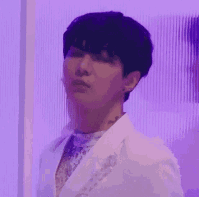 a man in a white suit is standing in front of a purple wall and looking at the camera .