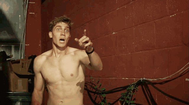 a man without a shirt is pointing at something in front of a red brick wall
