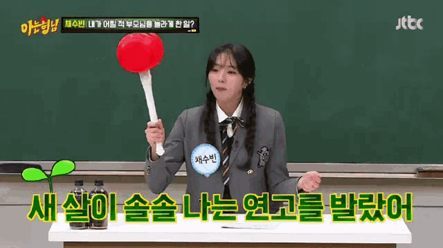 a girl in a school uniform is standing in front of a chalkboard holding a red object .