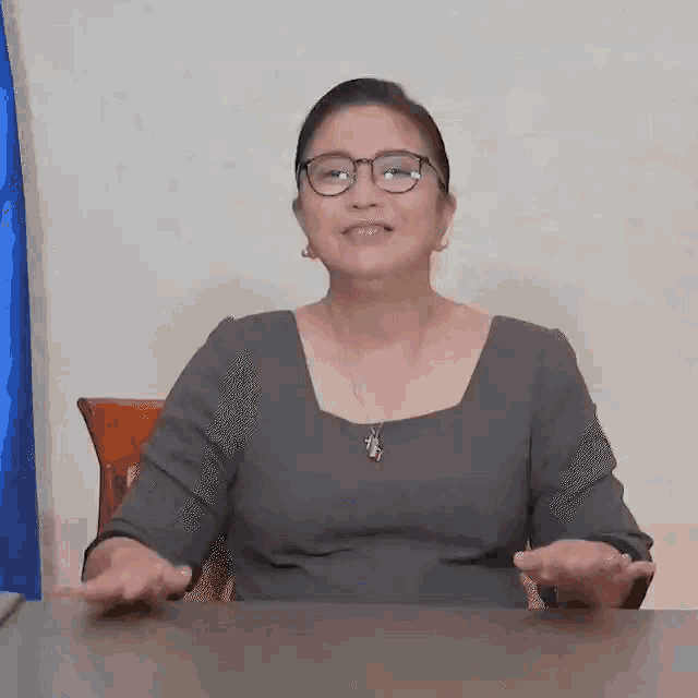 a woman wearing glasses and a cross necklace is sitting at a desk giving a thumbs up