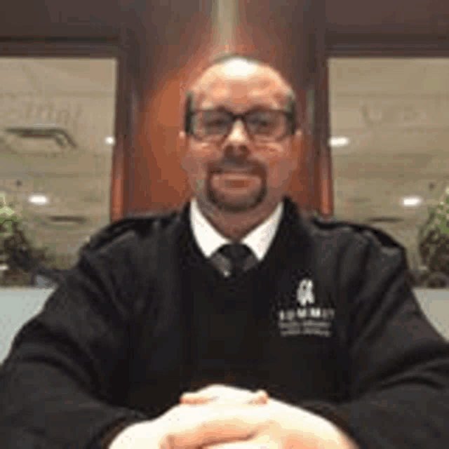a man with a beard and glasses is sitting at a table with his hands folded in front of a window .