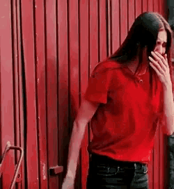 a woman in a red shirt is covering her mouth with her hand while standing in front of a red door .