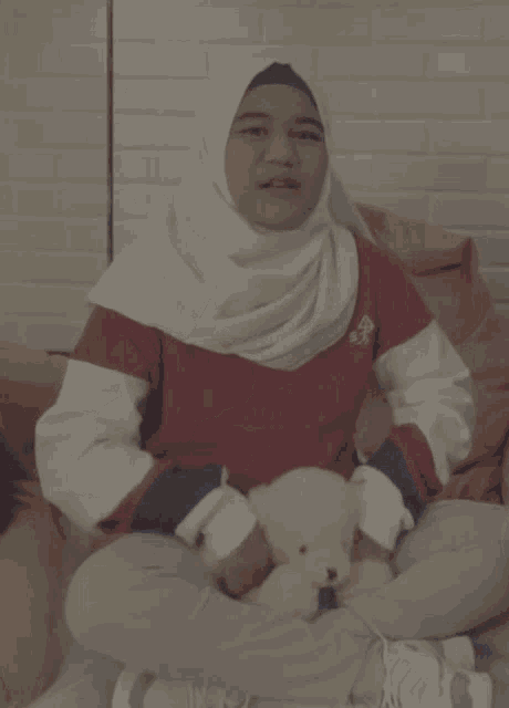 a woman wearing a hijab is sitting on a couch holding a teddy bear and giving a thumbs up