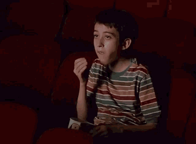 a young boy in a striped shirt is sitting in a movie theater eating popcorn