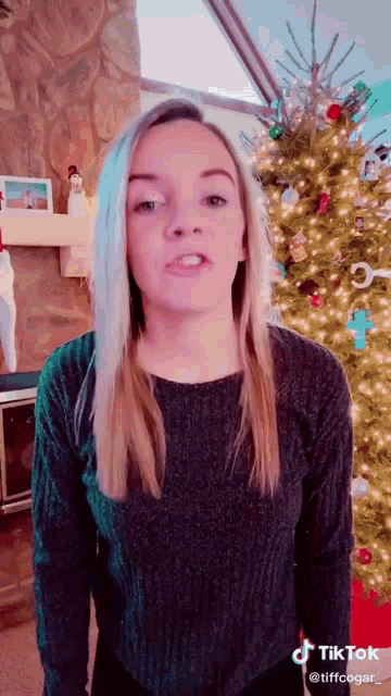 a woman in a black sweater is standing in front of a christmas tree and talking .