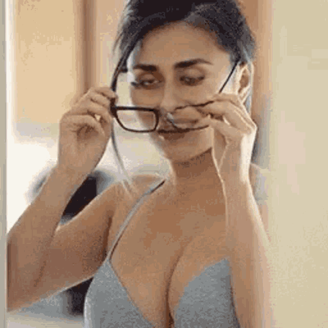 a woman in a bra is looking at herself in the mirror and putting on glasses .