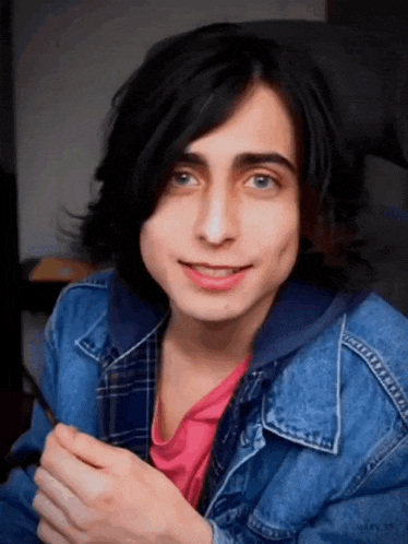 a young man with long black hair and blue eyes is wearing a blue denim jacket and a pink shirt .