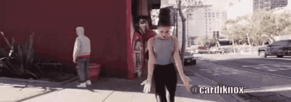 a woman is dancing on a sidewalk in front of a red building with the hashtag cardi knox .