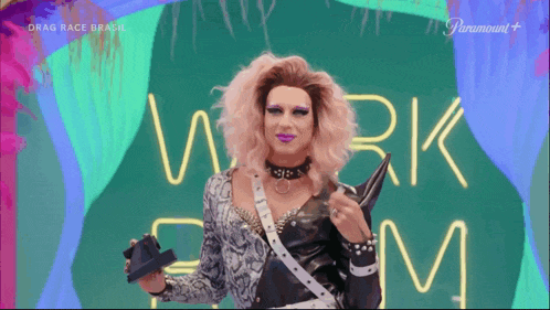 a drag queen stands in front of a neon sign that says work