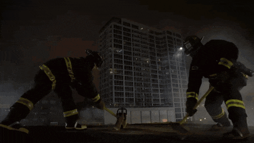 two firefighters in front of a building with a hammer and chainsaw