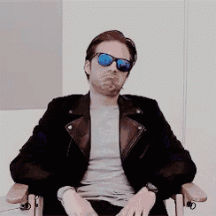 a man wearing sunglasses and a leather jacket is sitting in a chair and making a funny face .