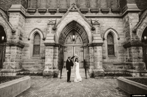 a black and white photo of a bride and groom standing in front of a brick building