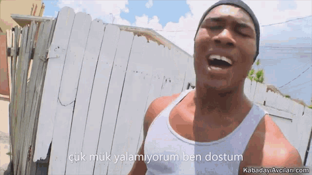 a man in a white tank top stands in front of a wooden fence with kabadayiavcian.com written on the bottom
